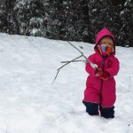 Ein kleines Kind in rotem Overall und Schal ums Gesicht steht mit Stecken in der Hand im tiefen Schnee