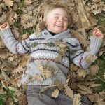 Kind liegt mit Laub bedeckt auf der Wiese und freut sich