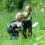 Zwei Kinder in Latzhose spielen mit Holzstecken im hohen Gras