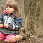 Mädchen kniet am erdigen Waldboden und blickt in die Ferne